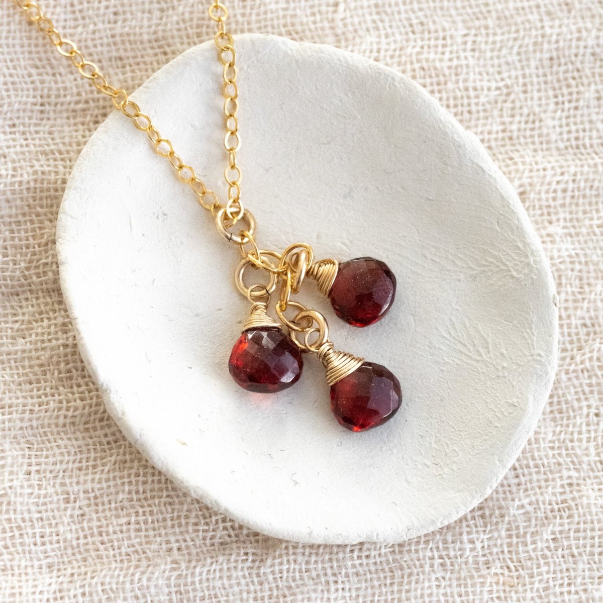 Eine feine Goldkette mit drei untereinander hängenden Granat Edelsteinen liegt auf einem weißen Schälchen.