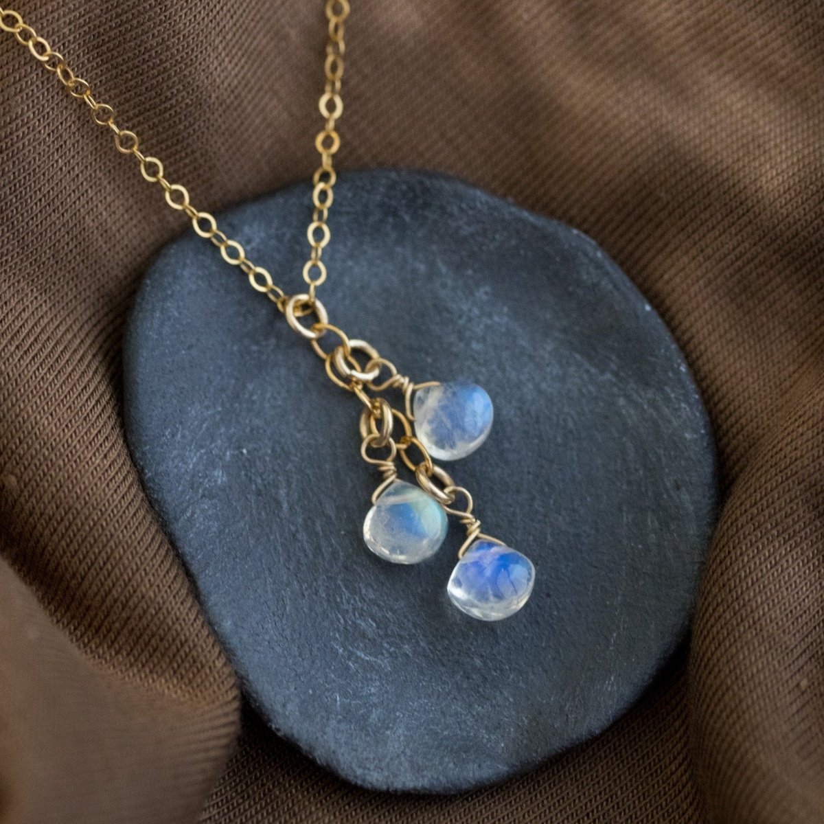 Eine feine Goldkette mit drei untereinander hängenden Mondsteintropfen liegt auf einem grauen Steinschälchen, das auf braunem Stoff steht.