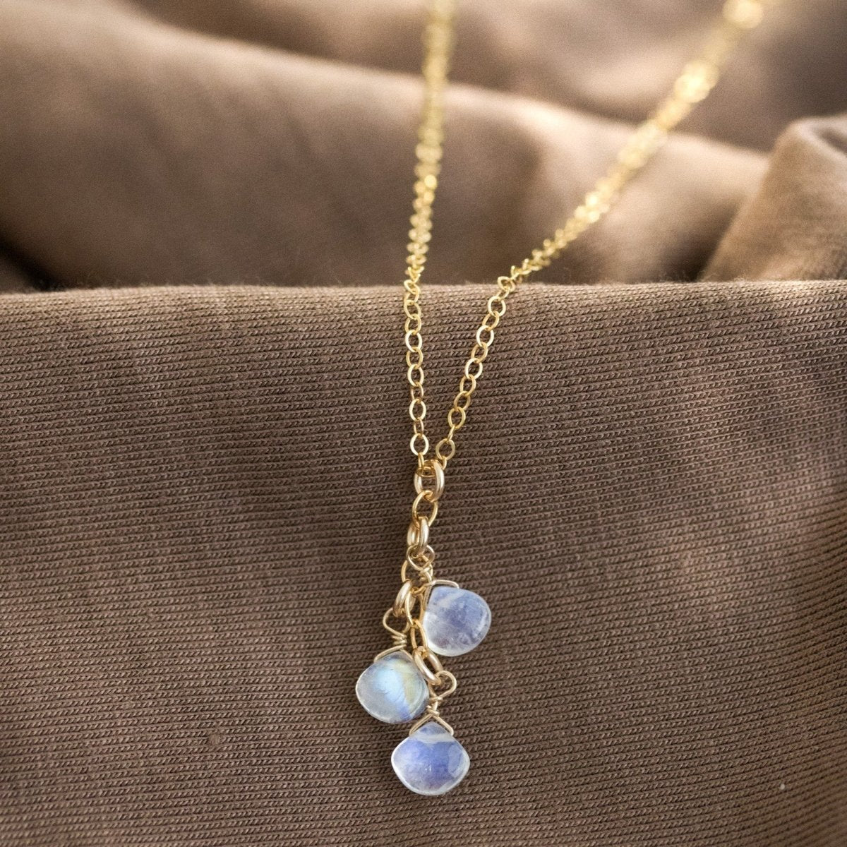 Eine feine Goldkette mit drei untereinander hängenden Mondsteintropfen lehnt an einem gewellten, braunen Stoff.