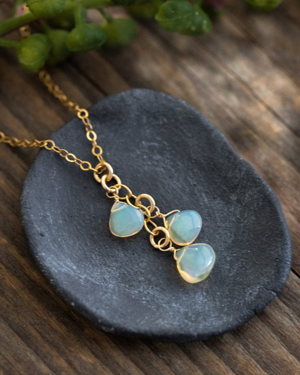 Eine feine Goldkette mit drei Opal-Anhänger liegt auf einem dunkelgrauen Schälchen, Holzuntergrund und verschwommener Grünzweig im Hintergrund.