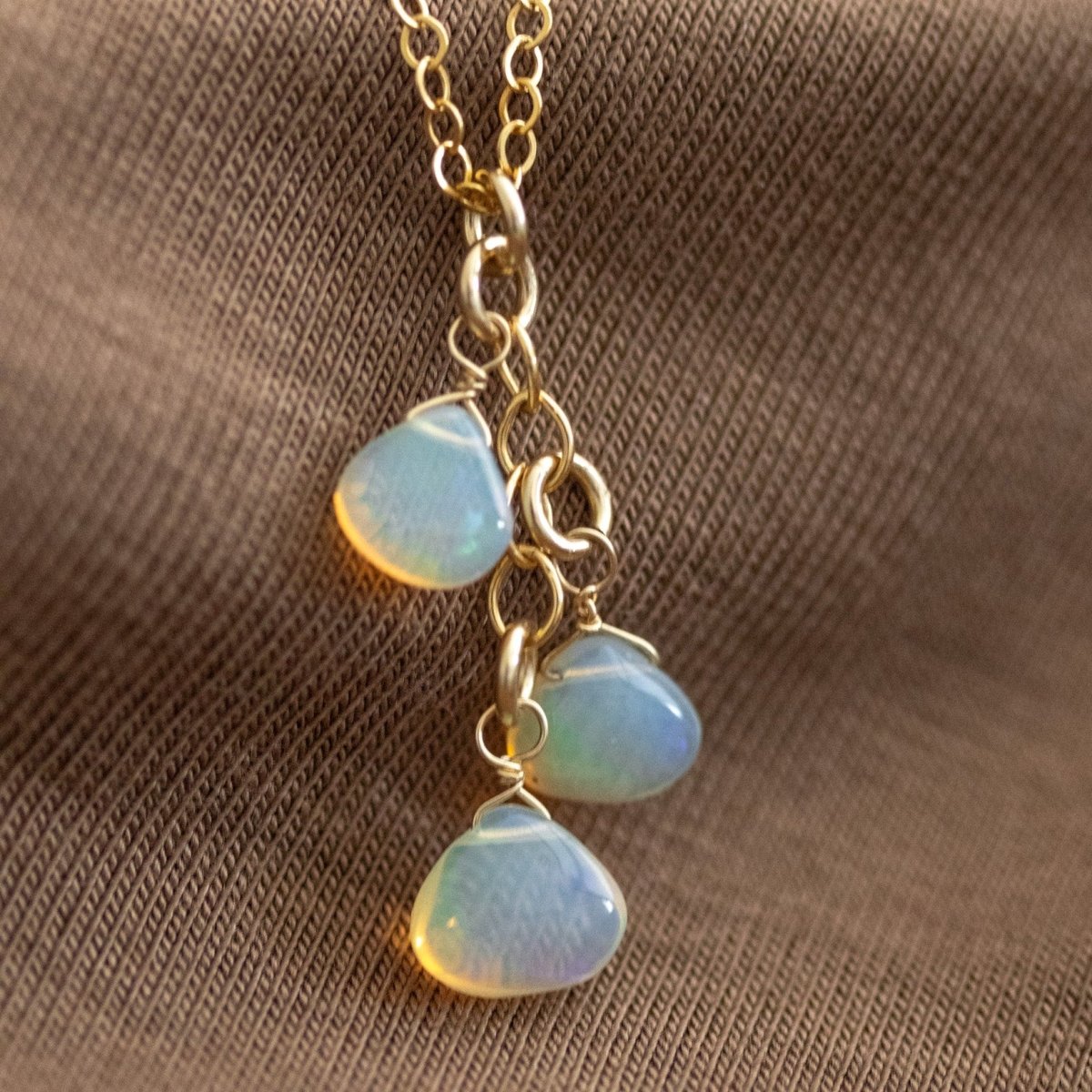 Eine feine Goldkette mit drei Opal-Anhänger liegt auf gewellten, braunen Stoff.