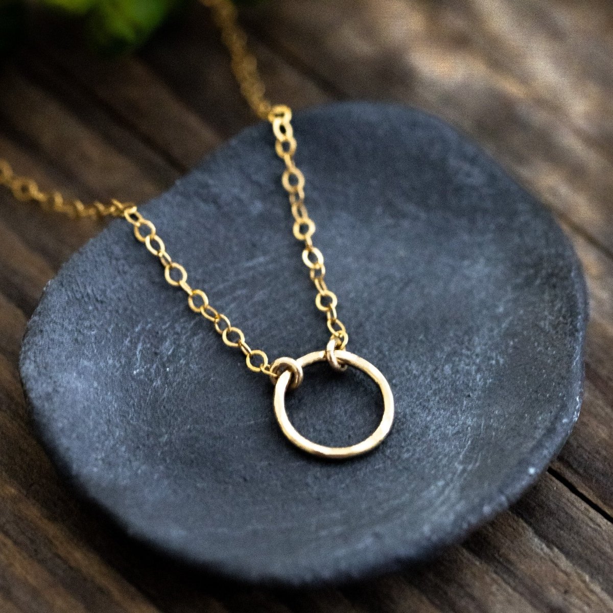 Eine feine, goldene Halskette mit einem Ring-Anhänger liegt auf einem grauen Steinschälchen, das auf Holzuntergrund steht.