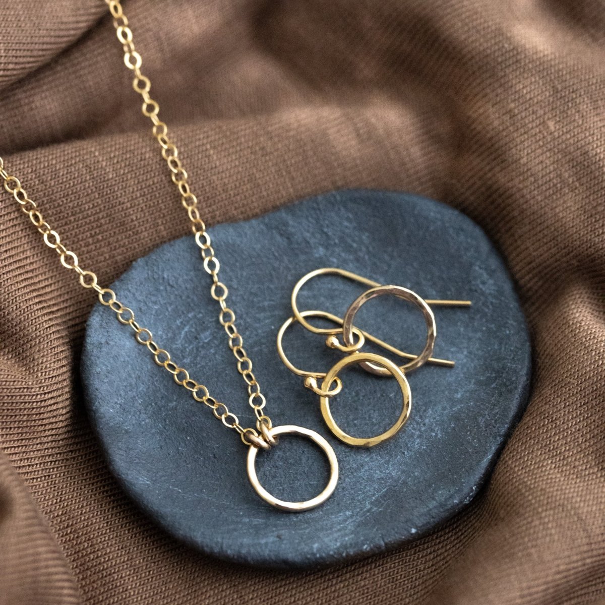 Ein Schmuckset, das aus einer feinen Goldkette mit Ring-Anhänger in der Mitte und einem Paar Ohrringen mit dem gleichen Ring besteht, liegt auf einer grauen Steinschale, die auf braunem Stoff liegt.