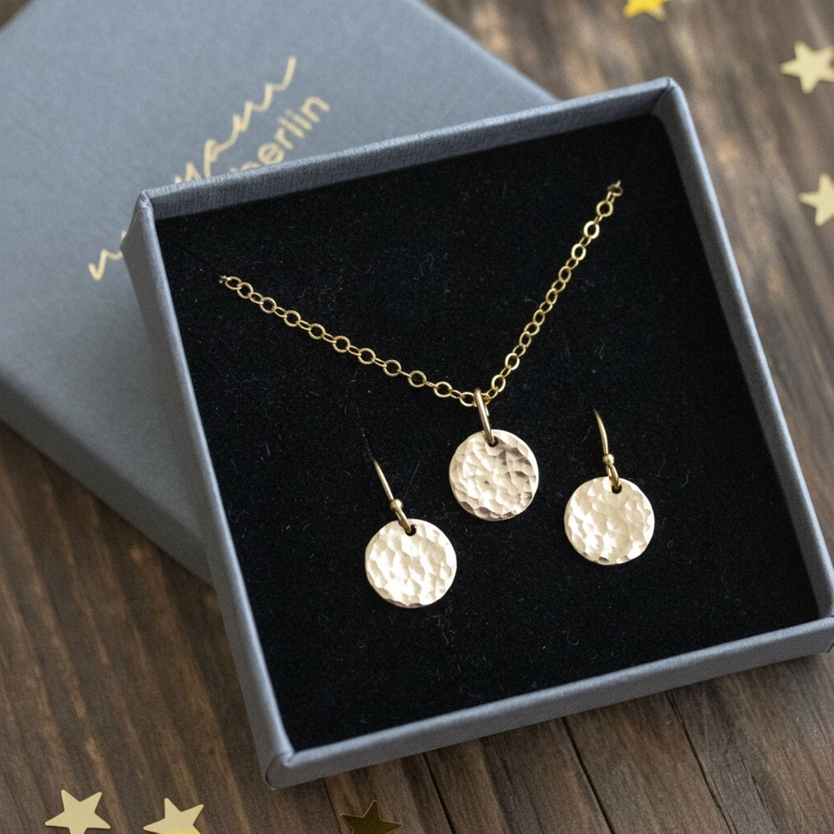 Ein goldenes Schmuckset ist in ein Schmucketui eingehängt. Es besteht aus einer feinen Kette und einem Paar Ohrringe, an denen jeweils ein gehämmertes Plättchen hängt. Auf dem Holzuntergrund liegen ein paar verstreute, kleine Konfetti-Sternchen.