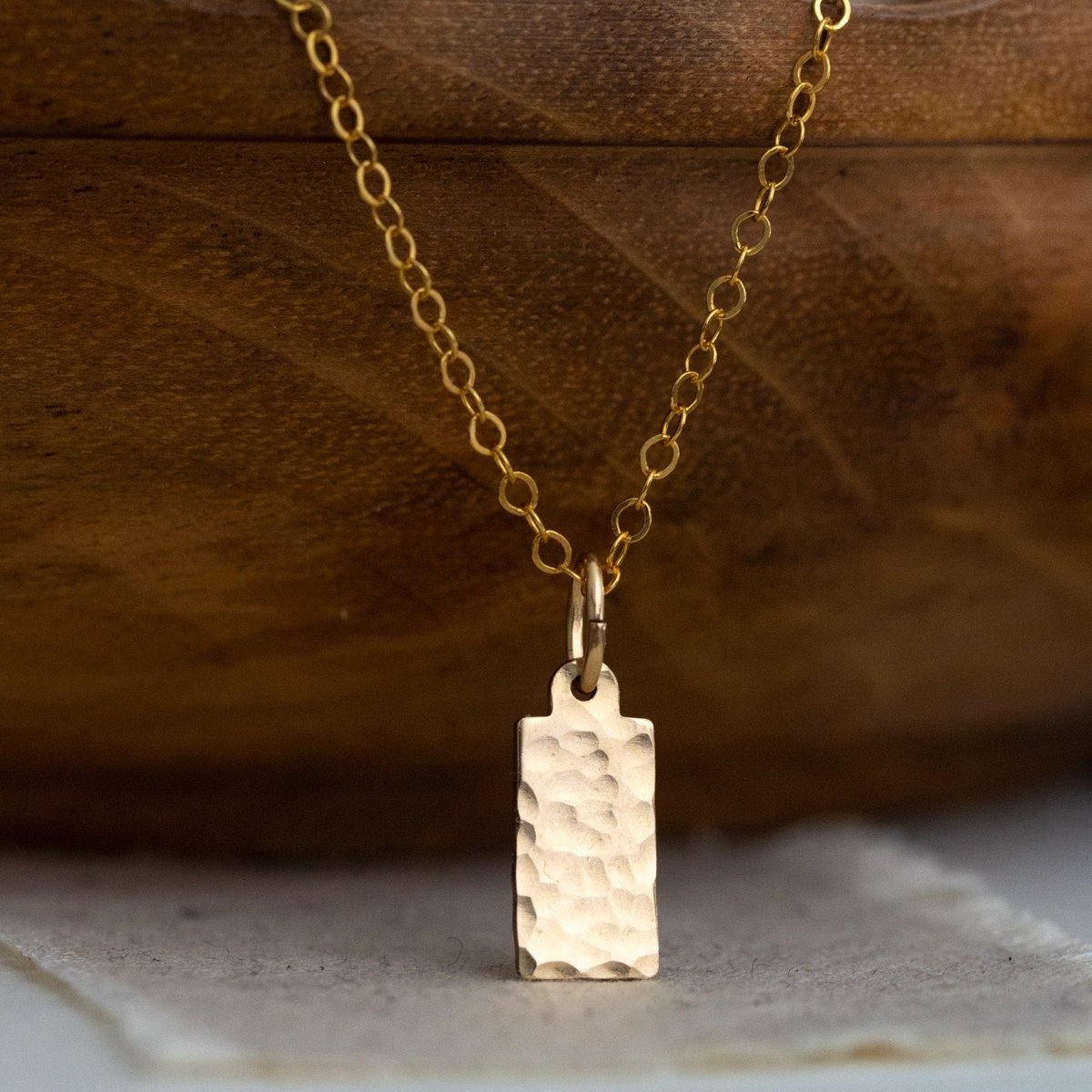 Eine feine und kurze Goldkette mit einem rechteckigen, gehämmerten Plättchen-Anhänger hängt von einer braunen, gewellten Holzschüssel herab.