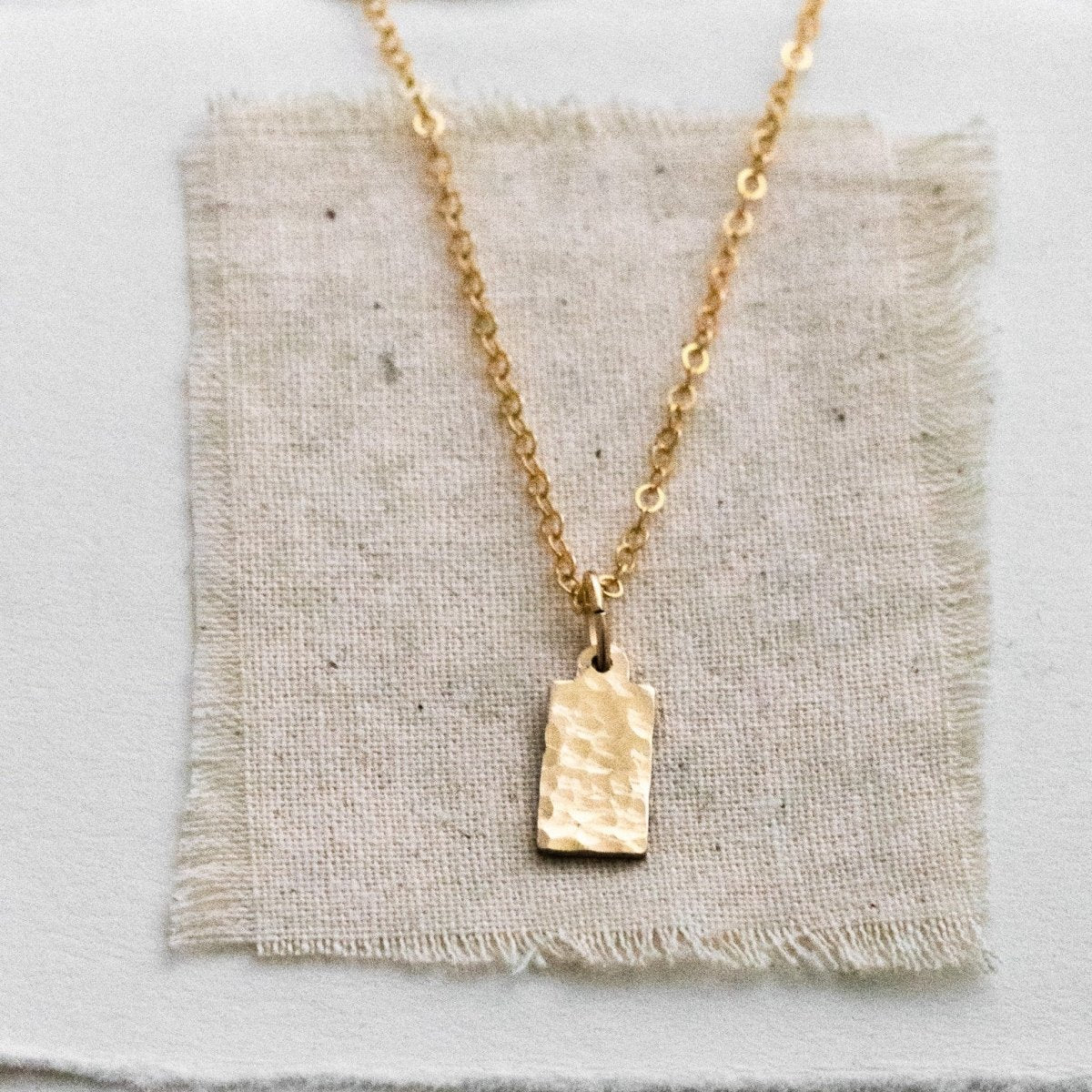 Eine feine und kurze Goldkette mit einem rechteckigen, gehämmerten Plättchen-Anhänger liegt auf einem naturfarbenen Leinenstoff.
