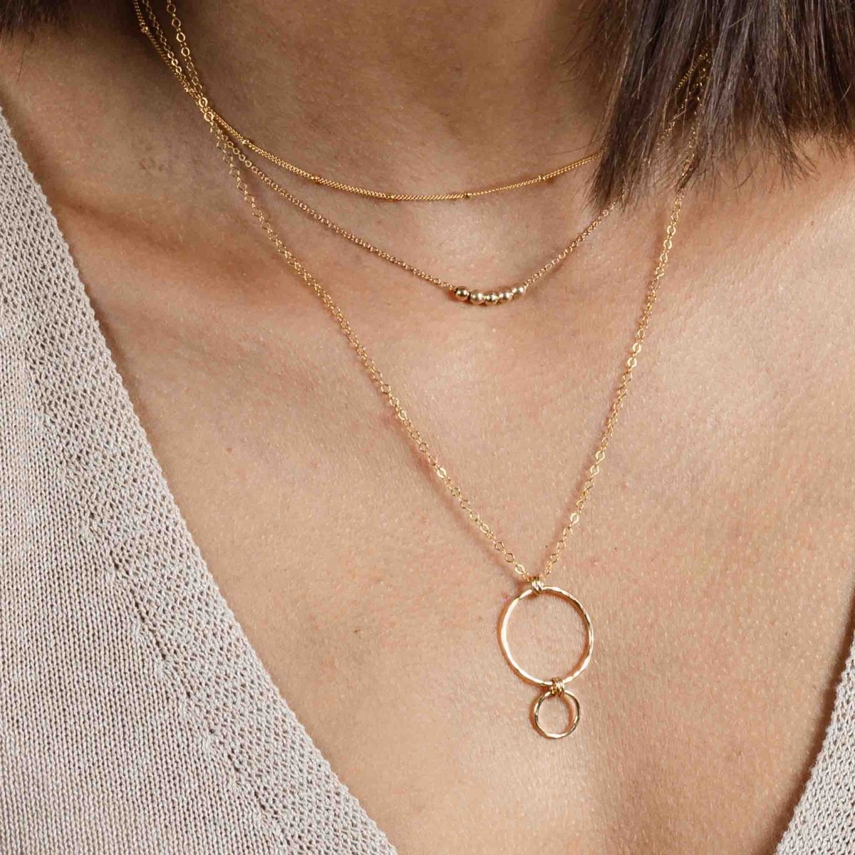 Am Hals des Models hängen drei Goldketten in verschiedenen Längen: eine ganz kurze Satellitenkette, eine feine Kette mit kleinen Goldperlen in der Mitte und eine längere Kette mit einem Anhänger, der aus gehämmerten Ringen besteht.