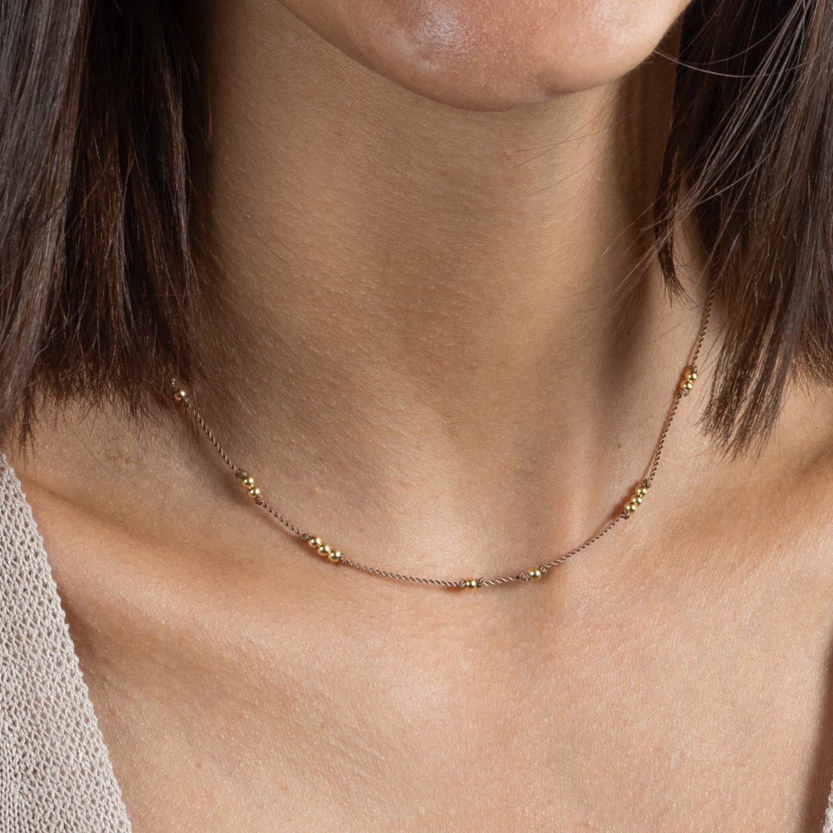 Eine dünne, beige Kordelkette mit unregelmäßig verteilten Goldperlen am Hals getragen.