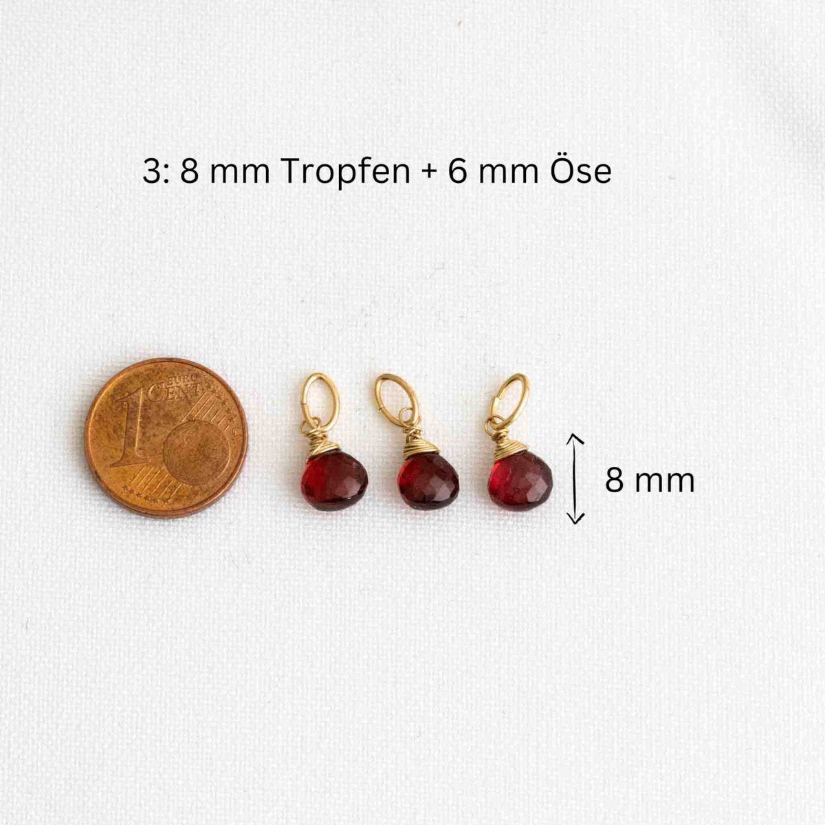 Drei kleine Granatanhänger neben einer 1-Cent-Münze auf weißem Hintergrund.