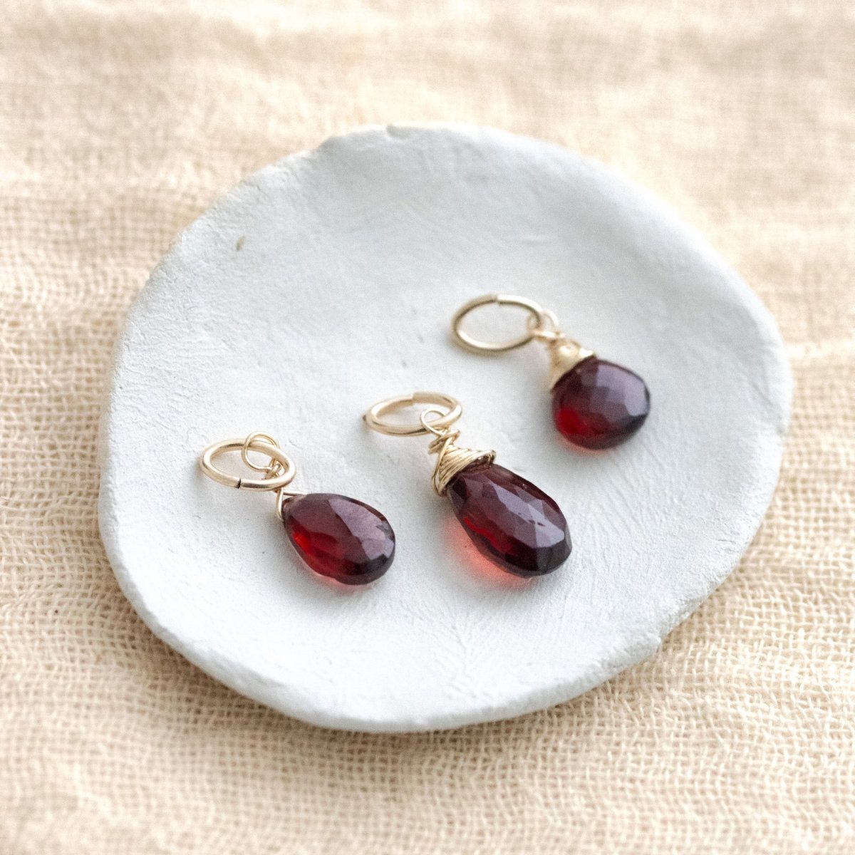 Drei gold gefasste Granatanhänger liegen auf einem weißen Schälchen auf hellem Leinenstoff.