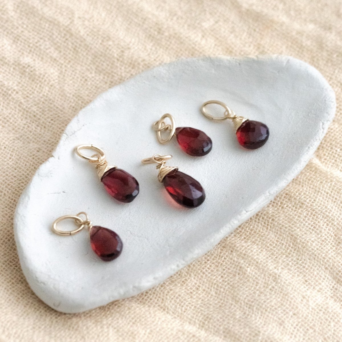 Fünf gold gefasste Granatanhänger liegen auf einem weißen Schälchen auf hellem Leinenstoff.