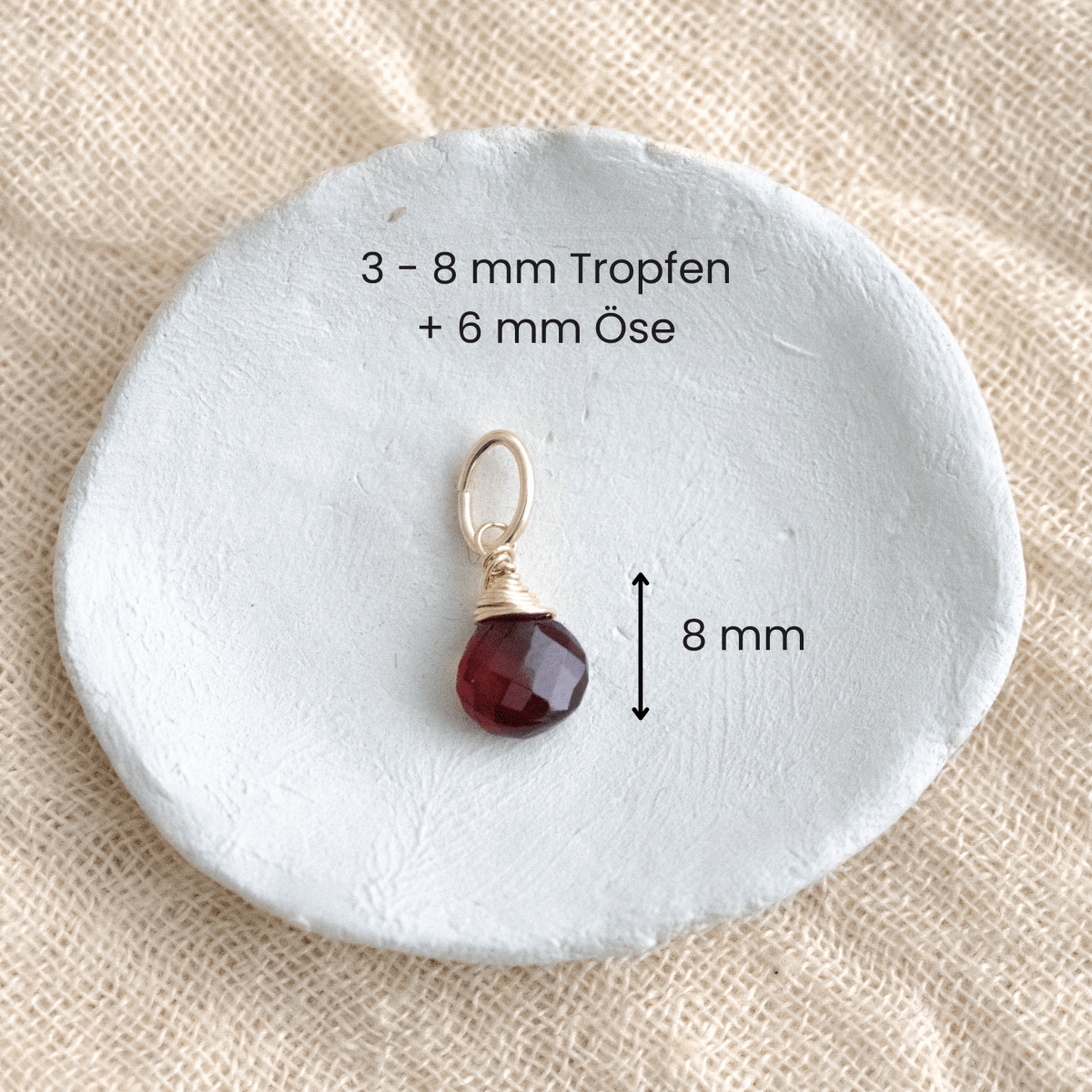 Ein Granat-Anhänger an goldener Öse liegen in einem weißen Schälchen auf hellem Stoff, mit Maßangaben.
