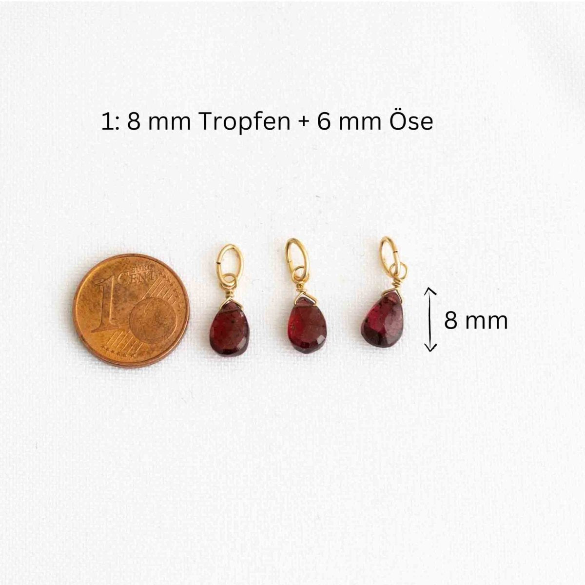Drei tropfenförmige Granatanhänger neben einer 1-Cent-Münze auf weißem Hintergrund.
