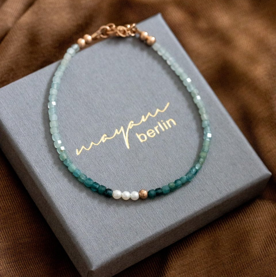 Ein Grandidierit-Edelstein-Armband mit Farbverlauf und weißen Süßwasserperlen in der Mitte liegt auf einem grauen Schmucketui.