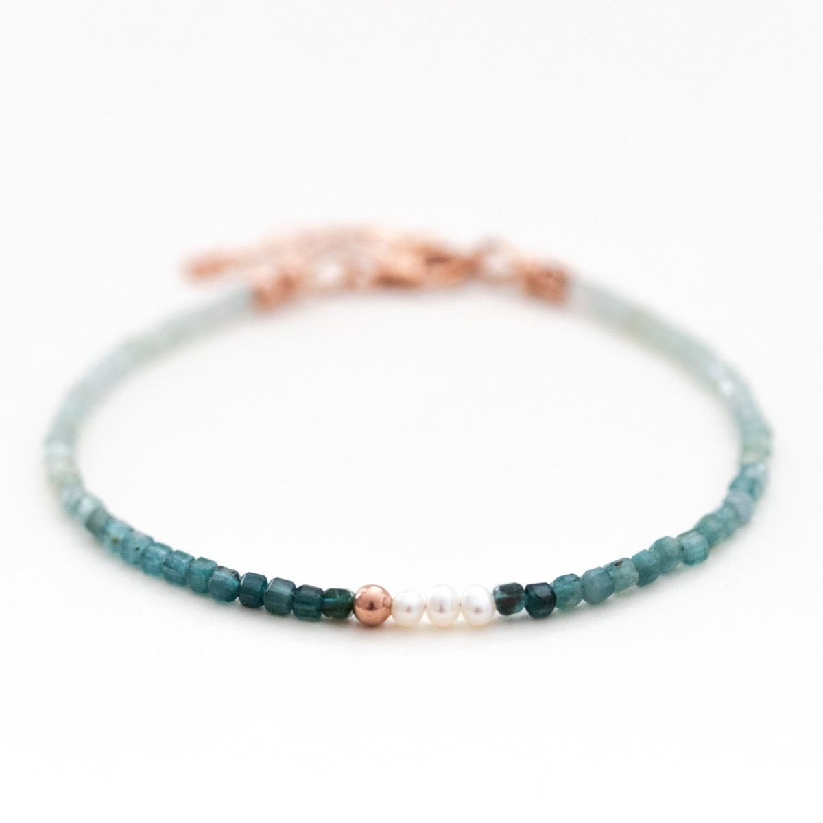 Ein dünnes Armband, bestehend aus kleinen Grandidierit-Perlen mit Farbverlauf und drei Süßwasserperlen in der Mitte liegt auf weißem Hintergrund.