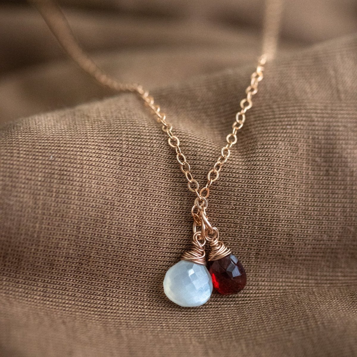 Ein dunkelroter Granat und ein grauer Mondstein an einer feinen Rosegold-Kette lehnen an braunem Stoff.