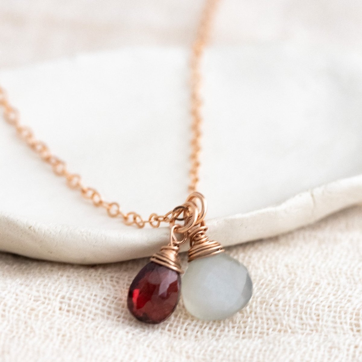Ein dunkelroter Granat und ein grauer Mondstein an einer feinen Rosegold-Kette lehnen an einem weißen Schälchen.
