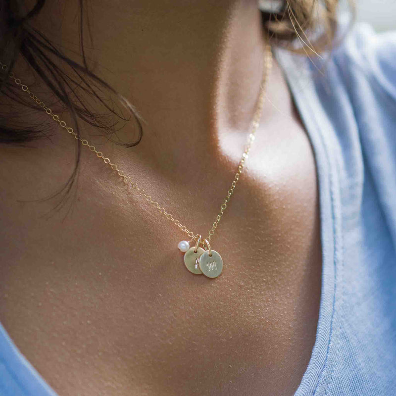 Am Hals des Models hängt eine kurze, goldene Kette mit einer kleinen Süßwasserperle und zwei Plättchen, bestempelt mit Initialen.