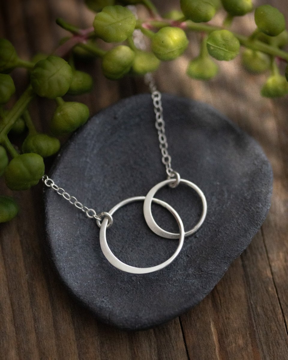 Eine zierliche 925 Silberkette mit zwei miteinander verschlungenen und unterschiedlich großen Ringen in der Mitte lehnt an einem grauen Schälchen auf Holzuntergrund. Ein verschwommener, grüner Zweig im Hintergrund.