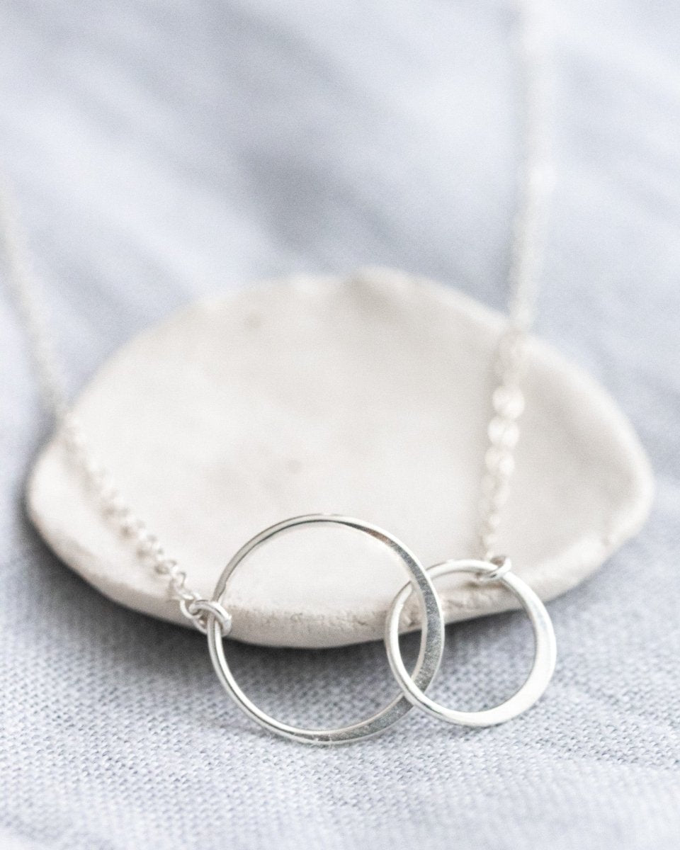 Eine zierliche 925 Silberkette mit zwei miteinander verschlungenen und unterschiedlich großen Ringen in der Mitte lehnt an einem weißen Schälchen.