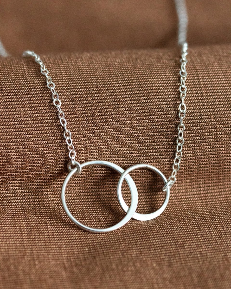 Eine zierliche 925 Silberkette mit zwei miteinander verschlungenen und unterschiedlich großen Ringen in der Mitte liegt auf braunem Stoff.
