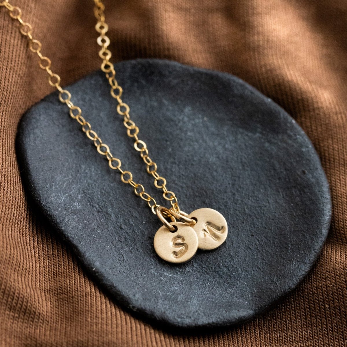 Eine feine Goldkette mit zwei runden Plättchen-Anhängern, auf die je ein Initial gestempelt wurde, liegt auf einem schwarzen Steinschälchen.