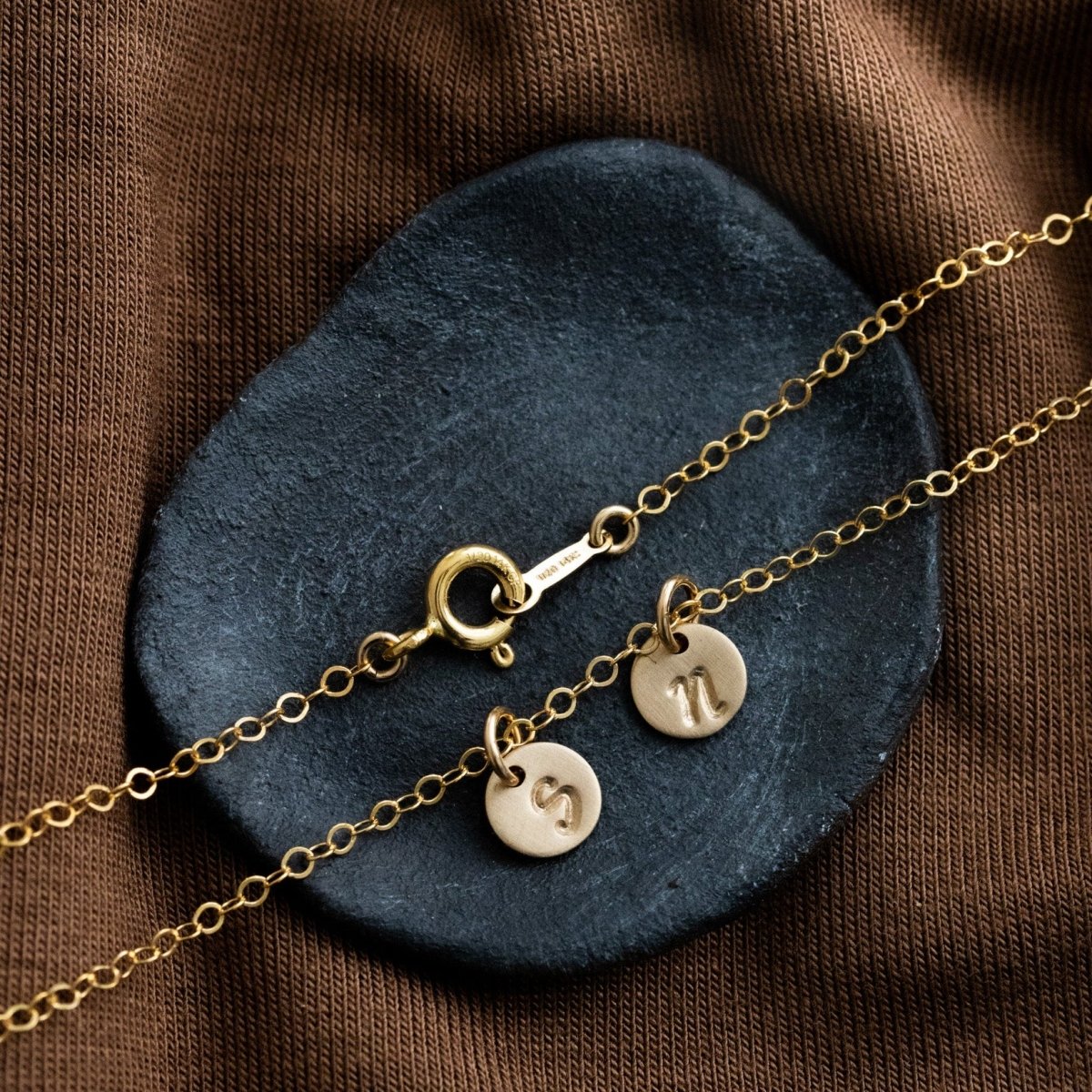 Eine feine Goldkette mit zwei runden Plättchen-Anhängern, auf die je ein Initial gestempelt wurde, liegt auf einem schwarzen Steinschälchen.