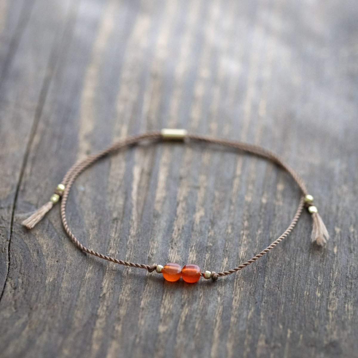 Dünnes Schnurarmband mit orangem Karneol und goldenen Akzenten auf Holz ausgelegt.