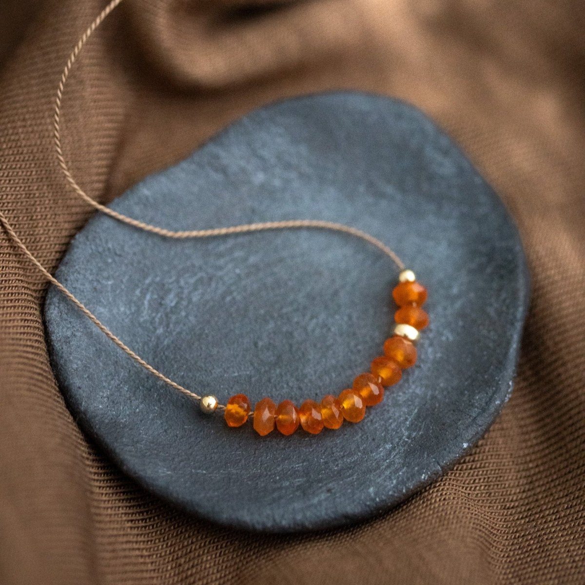 Auf einem dunkelgrauen Steinschälchen liegt eine dünne, beige Schnurkette, auf die eine Reihe oranger Karneolperlen und einige goldene Akzentperlen auf gezogen sind. Brauner Stoff als Untergrund.