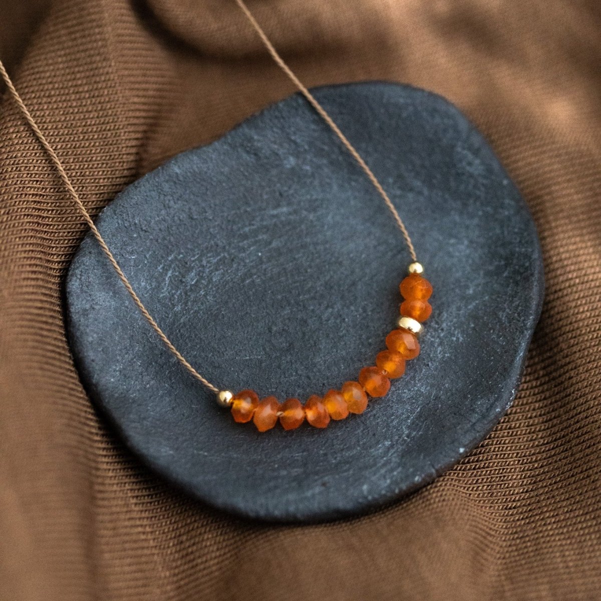 Auf einem dunkelgrauen Steinschälchen liegt eine dünne, beige Schnurkette, auf die eine Reihe oranger Karneolperlen und einige goldene Akzentperlen auf gezogen sind. Brauner Stoff als Untergrund.