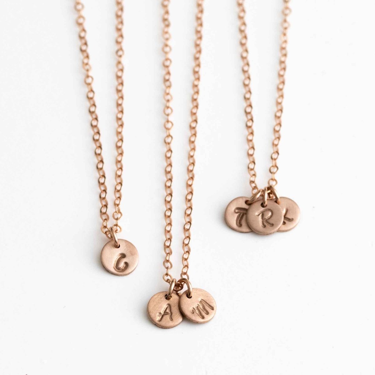 Drei Rosé Goldfill Ketten mit kleinen von Hand mit Buchstaben bestempelten Plättchen, hängen vor einem weißem Hintergrund.