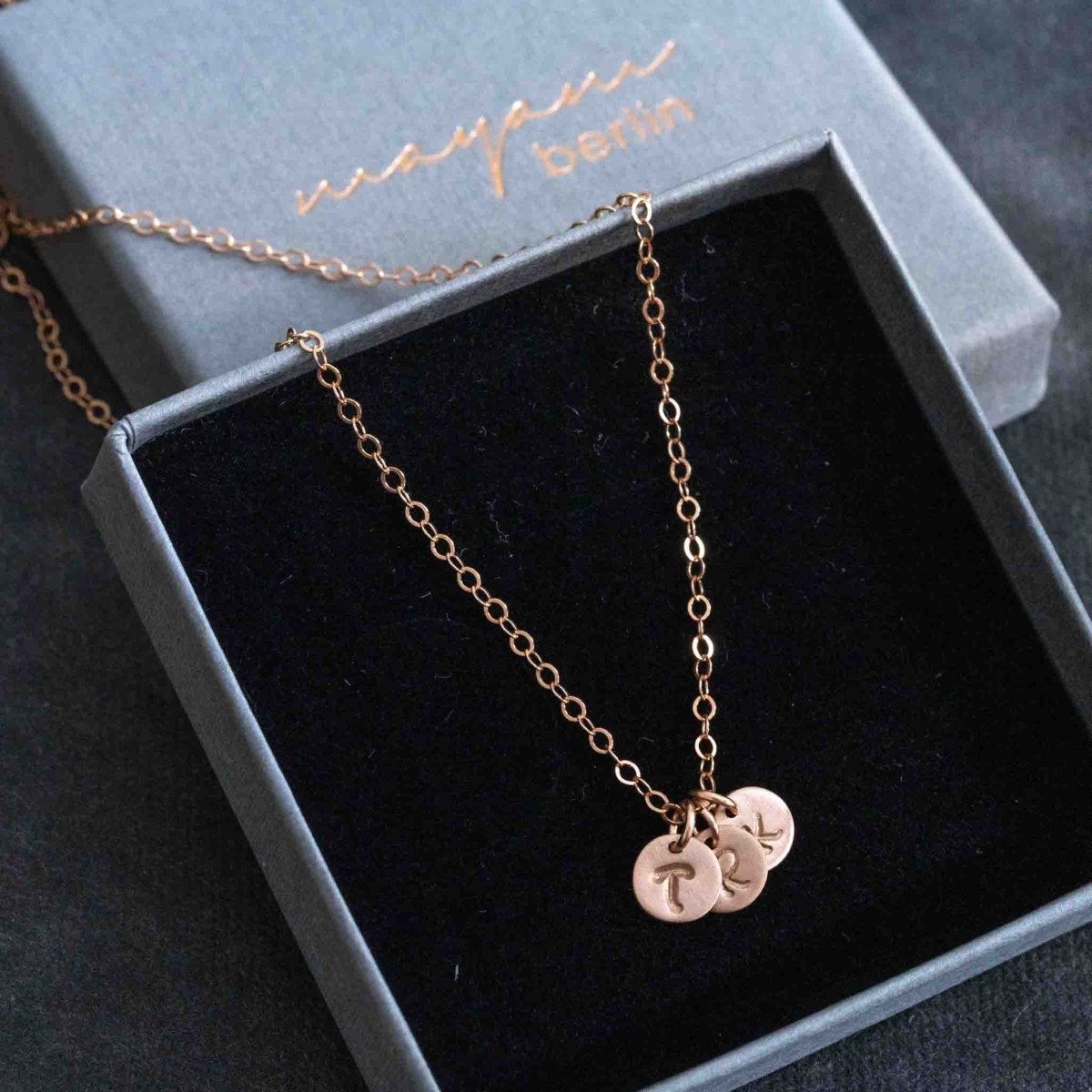 Eine rosé goldene Halskette mit drei runden Initial-Anhängern liegt auf einem grauen Schmucketui, in das der Name mayam berlin geprägt ist.