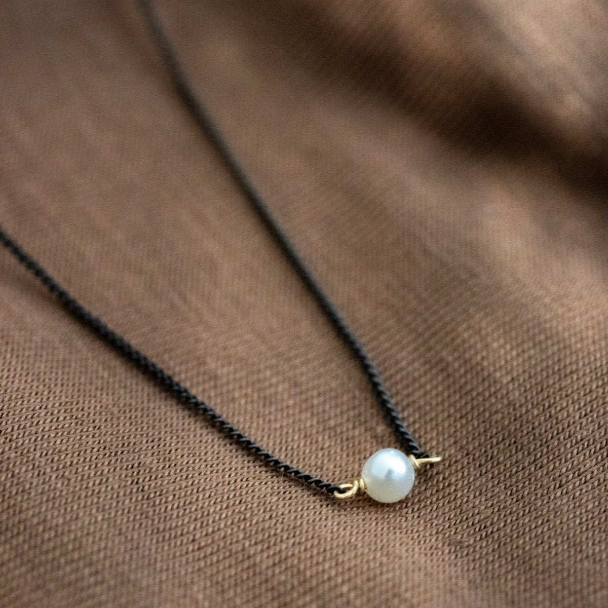 Eine dünne, schwarze Kette mit einer weißen Perle in der Mitte liegt auf braunem Stoff.