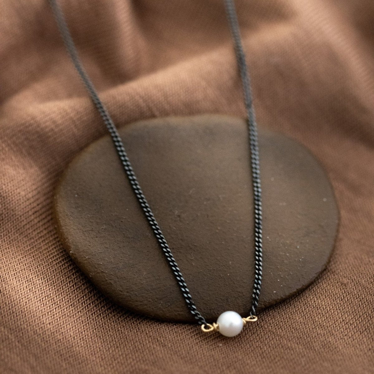 Eine dünne, schwarze Kette mit einer weißen Perle in der Mitte lehnt an einem braunen Schälchen auf braunem Stoff.
