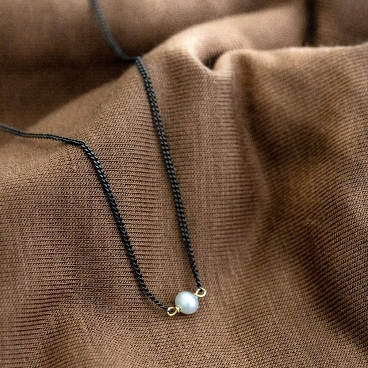Eine dünne, schwarze Kette mit einer weißen Perle in der Mitte hängt von braunem Stoff herunter.