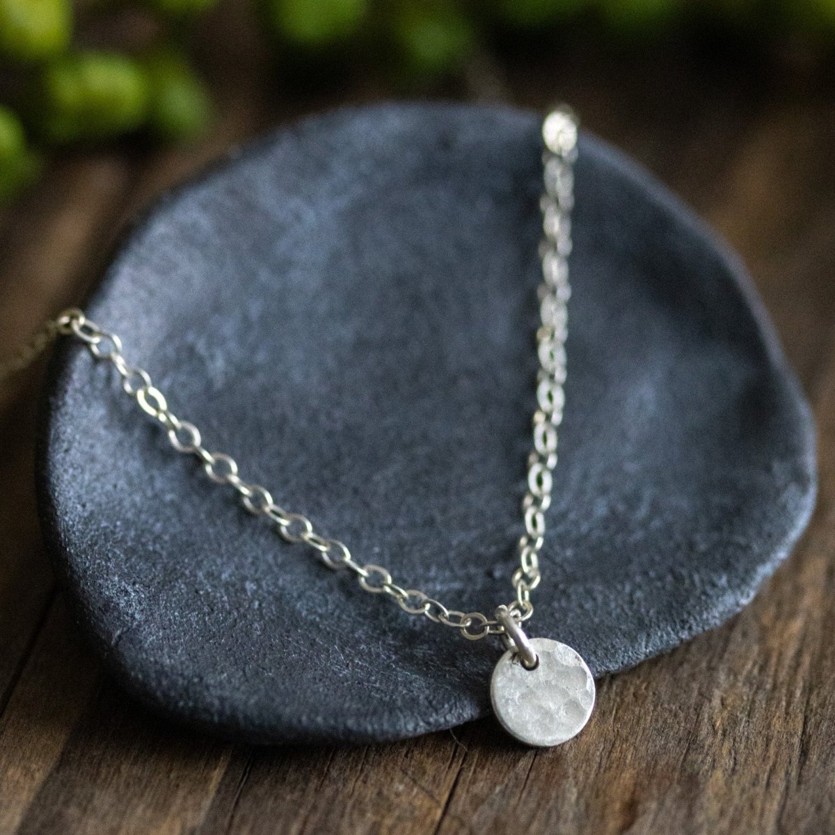 Eine feine 925 Silber-Ankerkette mit einem kleinen, runden Plättchen-Anhänger lehnt an einem grauen Schälchen auf Holzuntergrund. Im Hintergrund verschwommene Grünpflanzen.