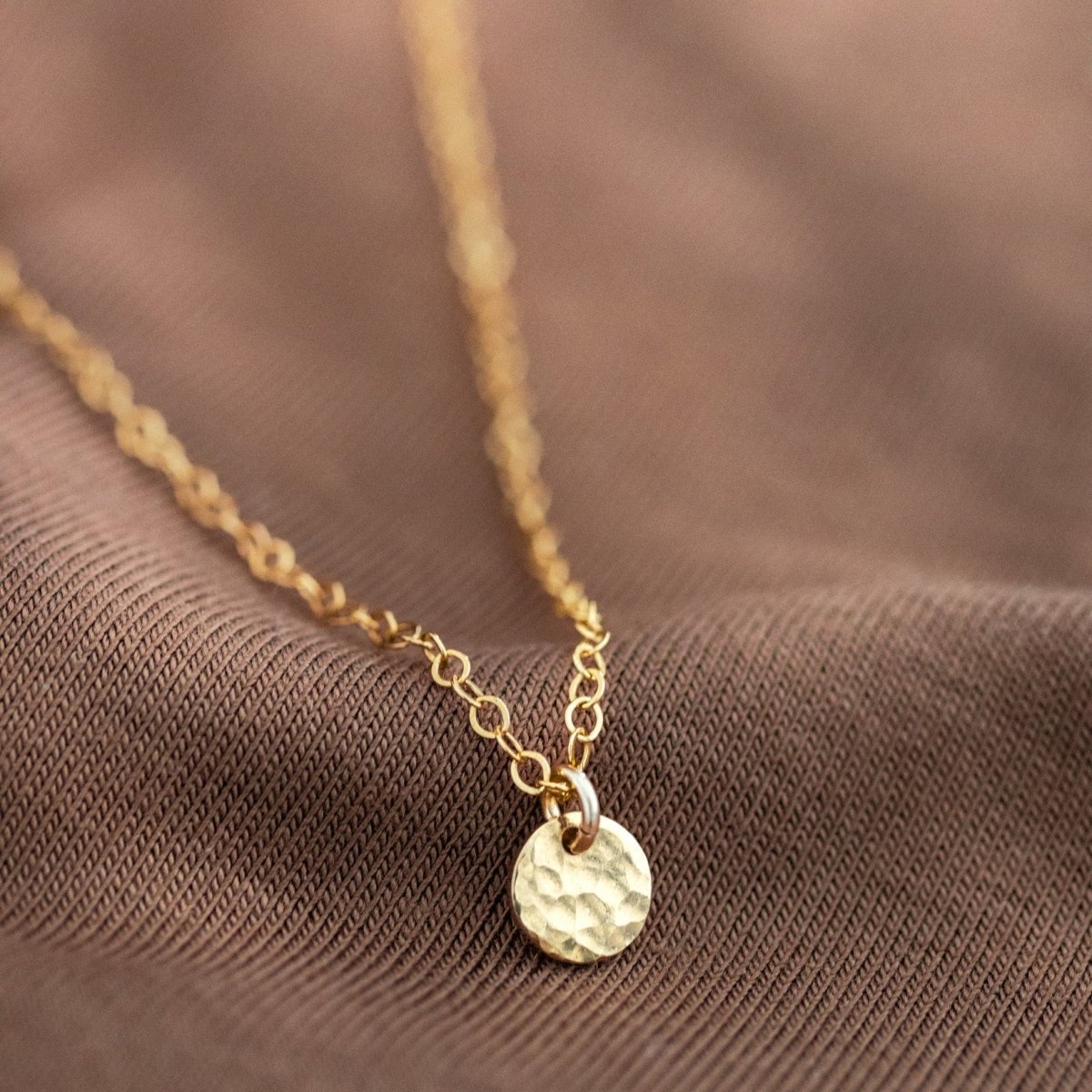 Eine goldene Ankerkette mit einem kleinen, gehämmerten Plättchen-Anhänger liegt auf braunem  Hintergrund.