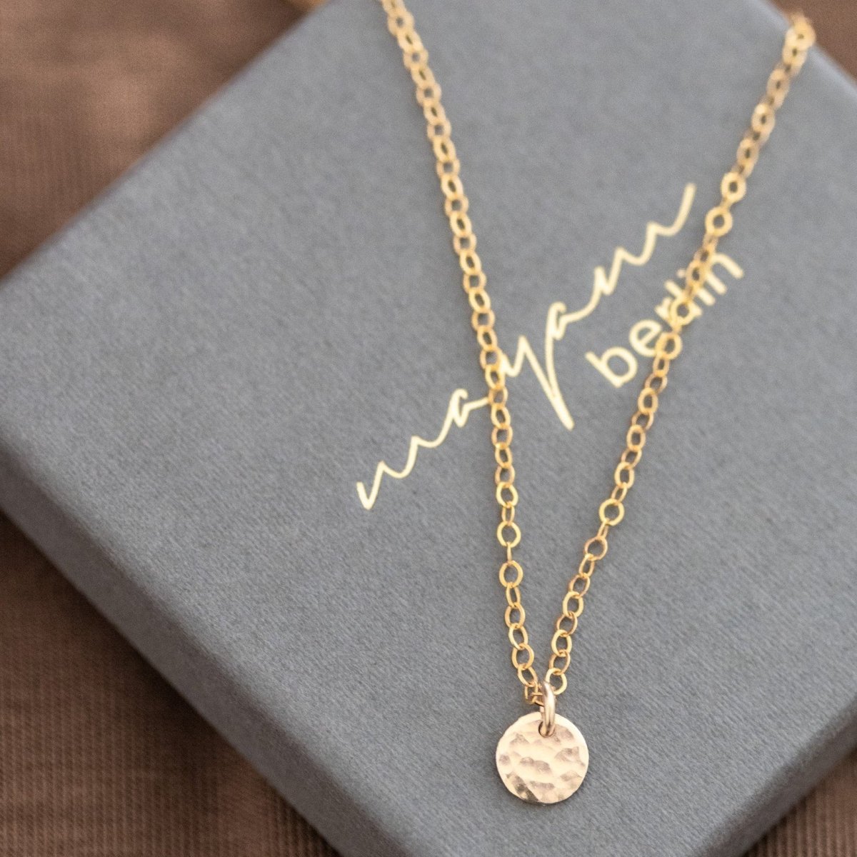 Eine goldene Ankerkette mit einem kleinen, gehämmerten Plättchen-Anhänger liegt auf einem grauen Schmucketui mit dem eingeprägten Namen mayam berlin