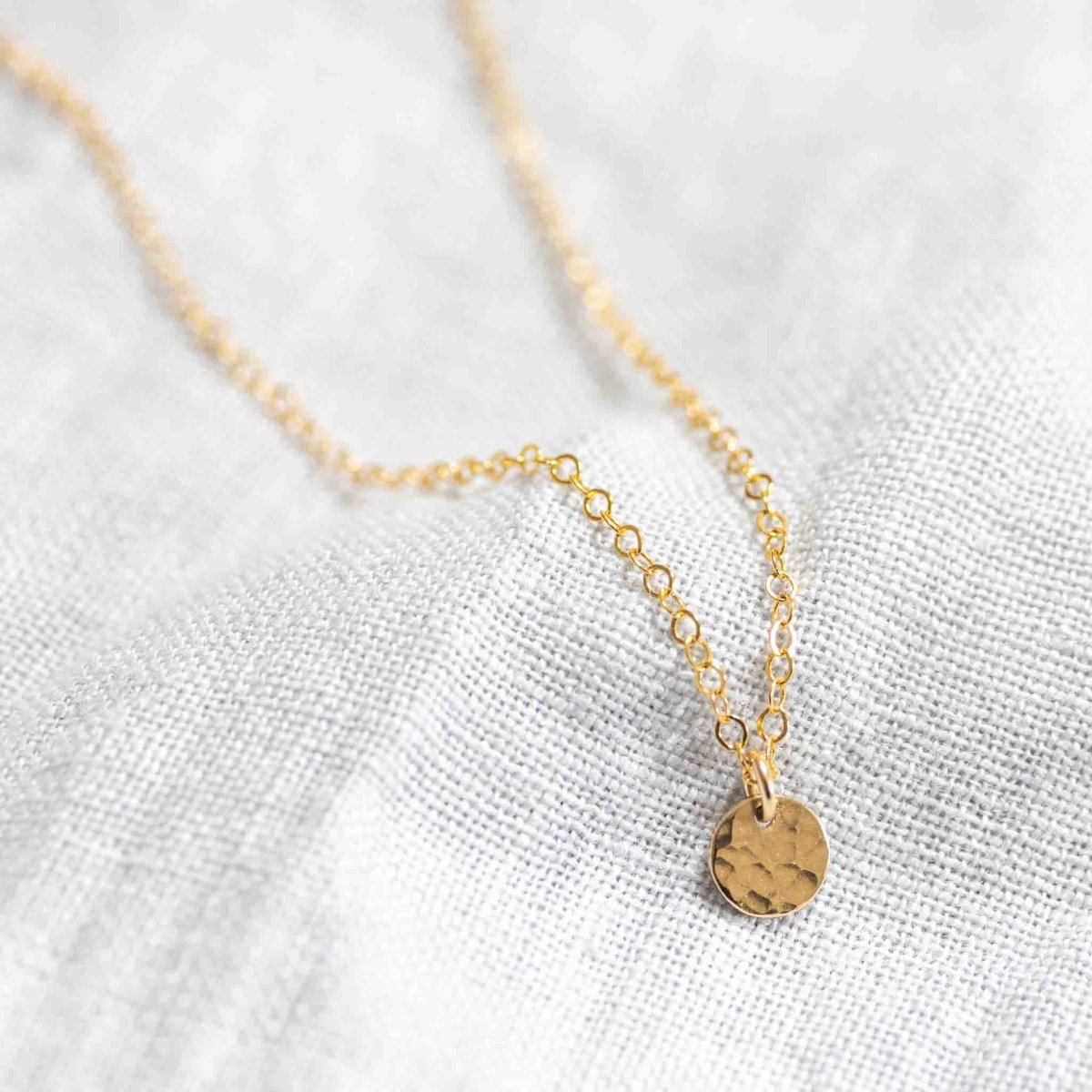 Eine goldene Ankerkette mit einem kleinen, gehämmerten Plättchen-Anhänger liegt auf weißem Leinen-Hintergrund.