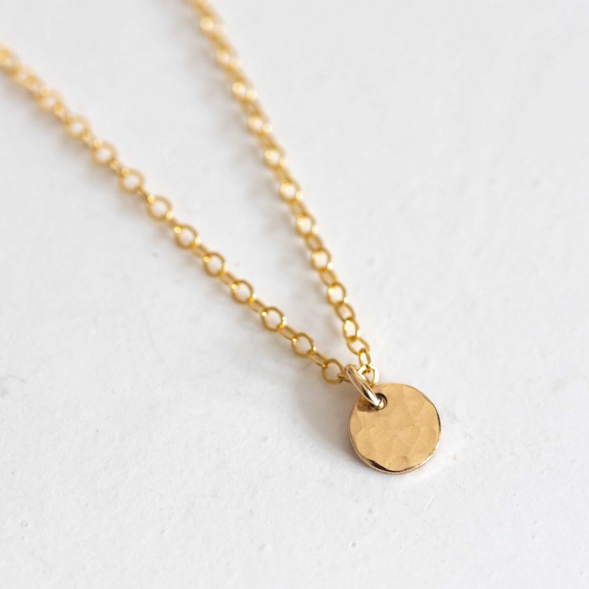 Eine goldene Ankerkette mit einem kleinen, gehämmerten Plättchen-Anhänger liegt auf weißem Hintergrund.