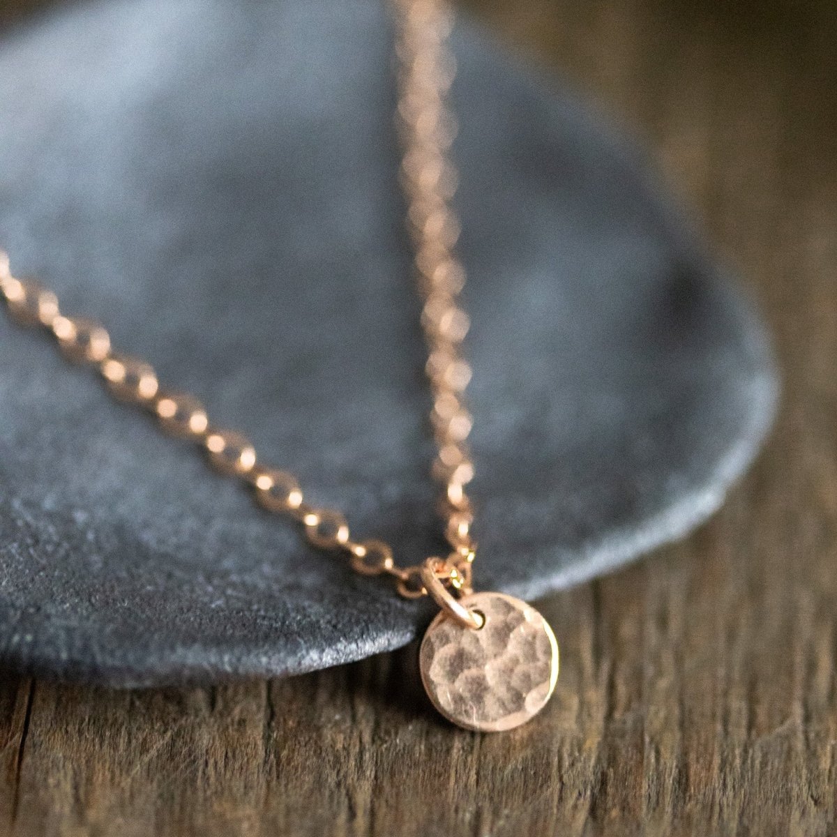 Eine feine rosé goldene Halskette mit einem kleinen, gehämmerten Plättchen lehnt an einem grauen Schälchen auf dunklem Holz. 