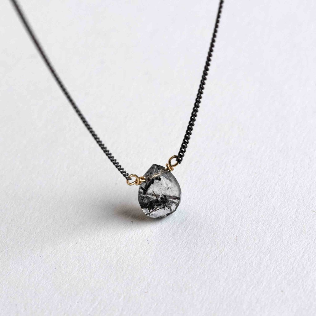Vor weißem Hintergrund hängt eine dünne, schwarze Panzerkette mit einem tropfenförmigen Rutilquarz Edelstein in der Mitte.