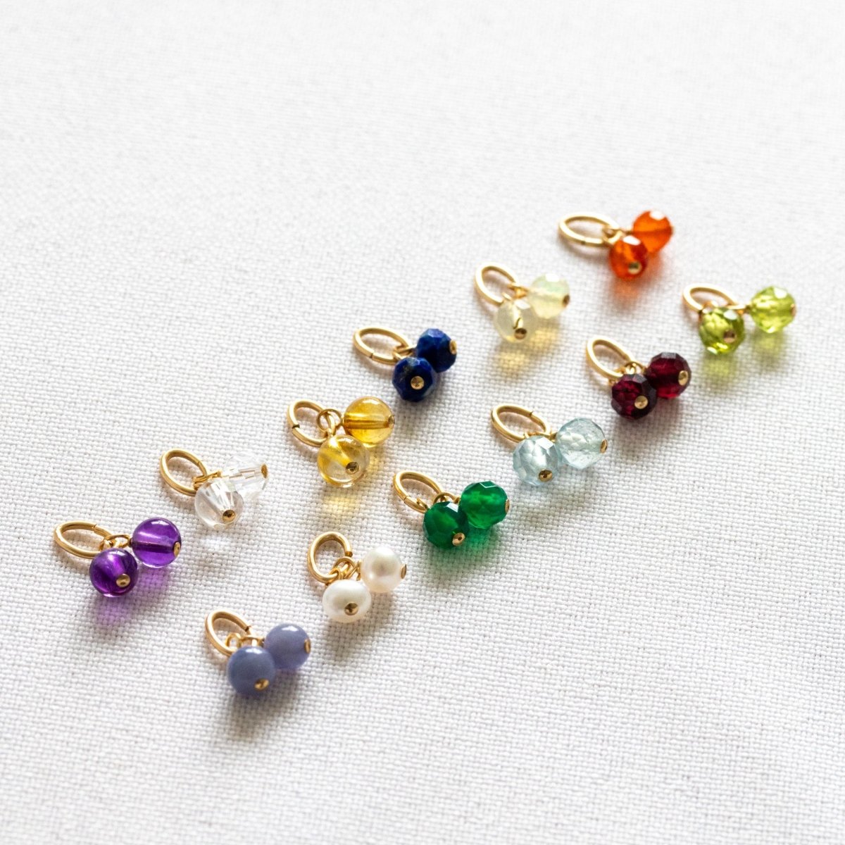 Zwei Reihen kleiner Geburtsstein-Anhänger liegen auf weißem Untergrund. Jeder Anhänger besteht aus zwei runden Edelsteinen, die an einer goldenen Öse hängen.