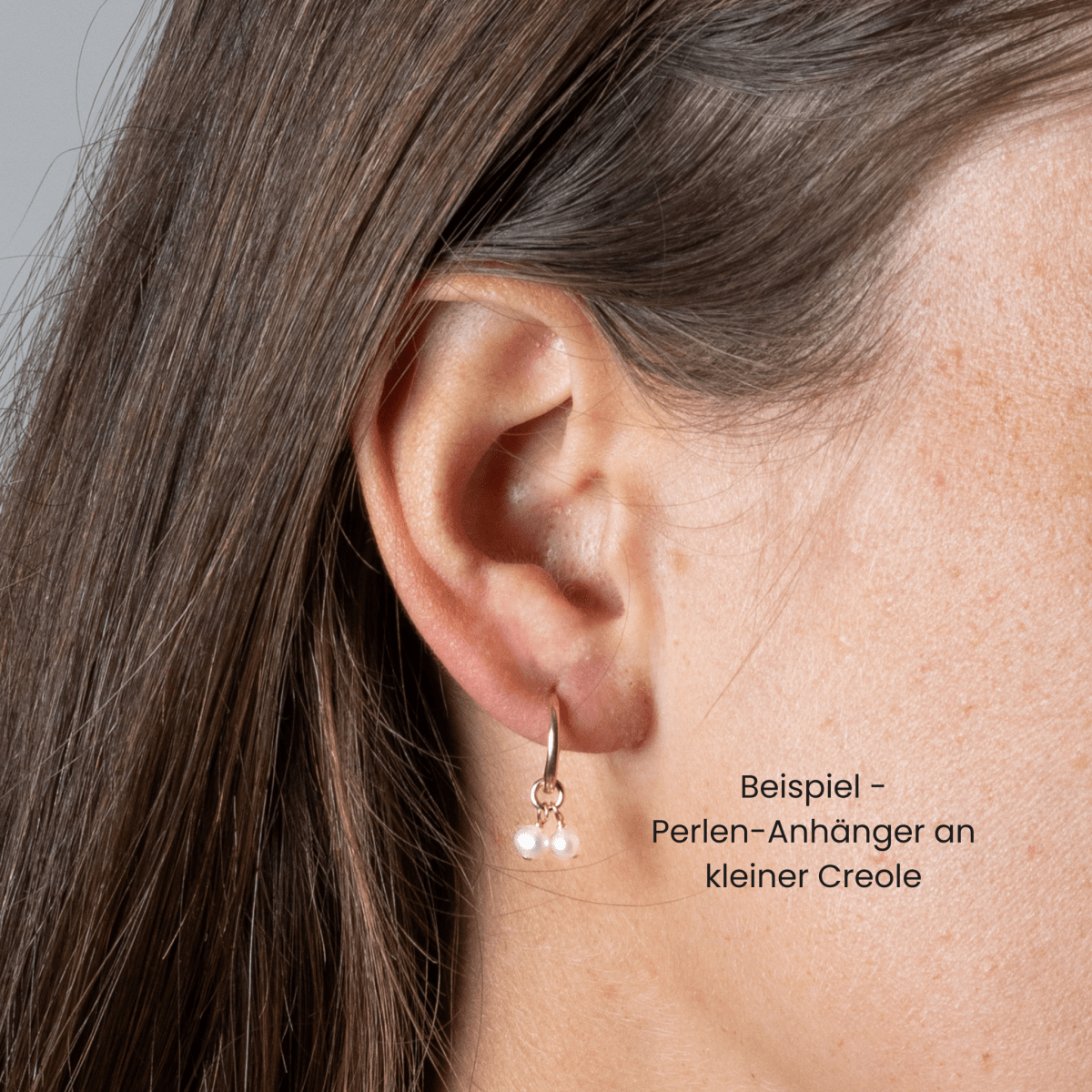 Am Ohr des Models hängt eine goldene Creole, an der zwei kleine, weiße Perlen baumeln. Daneben steht auf Deutsch: Beispiel - Perlen-Anhänger an kleiner Creole.