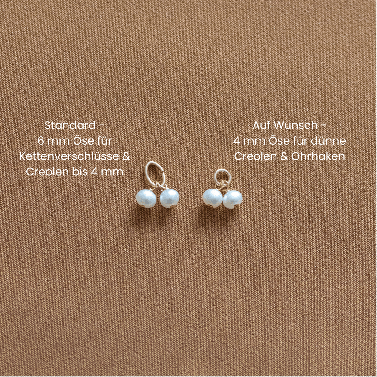 Zwei kleine weiße Perlen-Anhänger liegen mit unterschiedlich großen Ösen auf braunem Hintergrund. Daneben gibt es Hinweise zu den Ösen auf Deutsch.