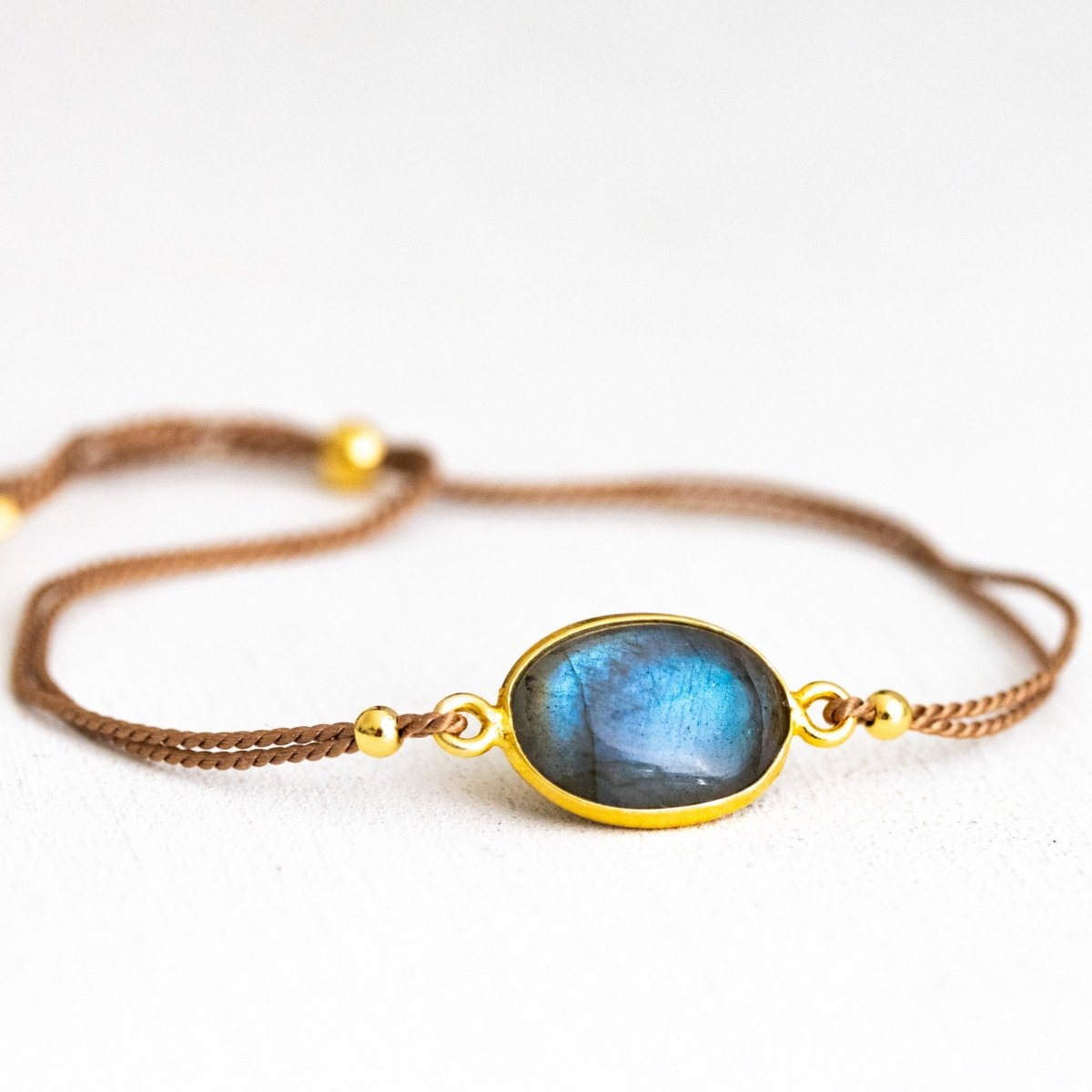 Ein dünnes, beiges Seidenarmband mit einem ovalen und in gold gefassten Labradorit in der Mitte liegt auf weißem Hintergrund.