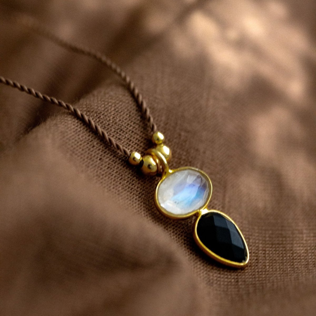 Eine dünne, braune Schnurkette mit einem Mondstein- und Onyx-Anhänger und goldenen Zierelementen liegt auf braunem Stoff.