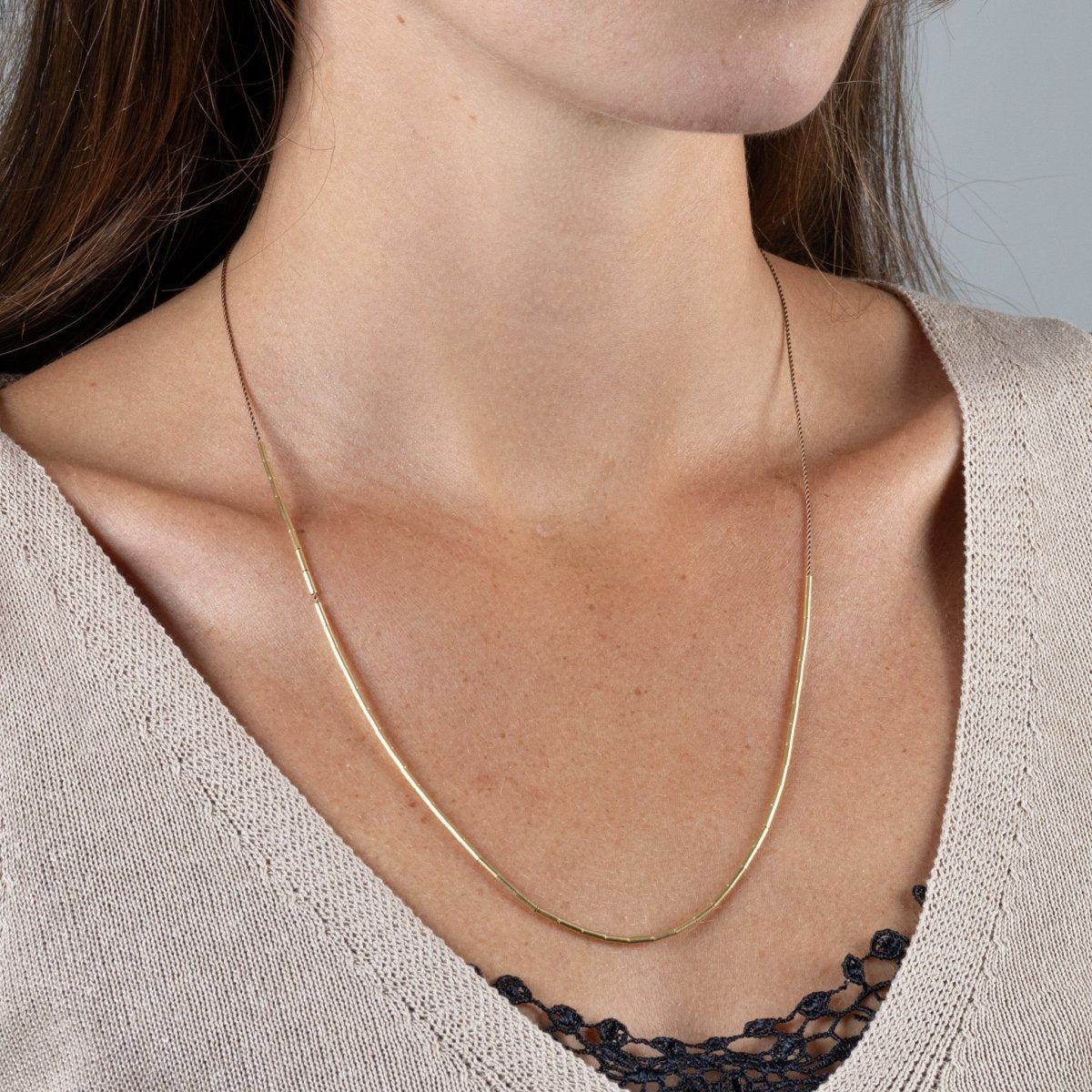 Eine zierliche Schnurkette mit einer fließenden Linie aus schmalen Goldröhrchen, getragen von einer Frau in einem beigefarbenen V-Ausschnitt-Pullover.
