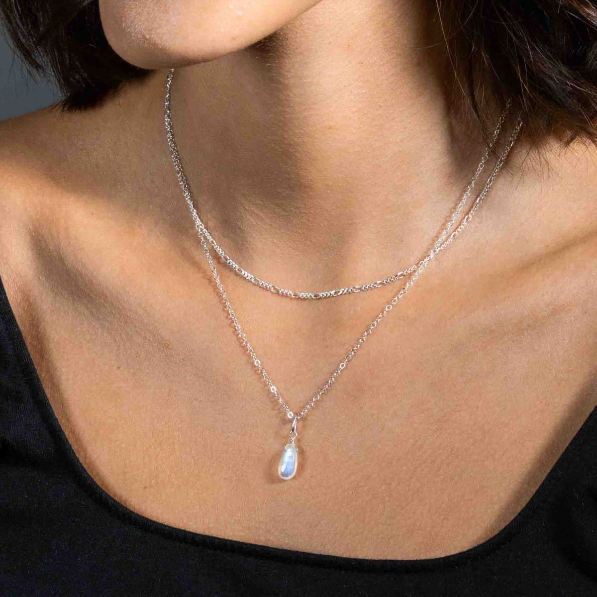 Am Hals des Models hängt eine kürzere Figaro-Kette aus Silber und eine längere Ankerkette aus Silber mit einem tropfenförmigen Mondstein-Anhänger.