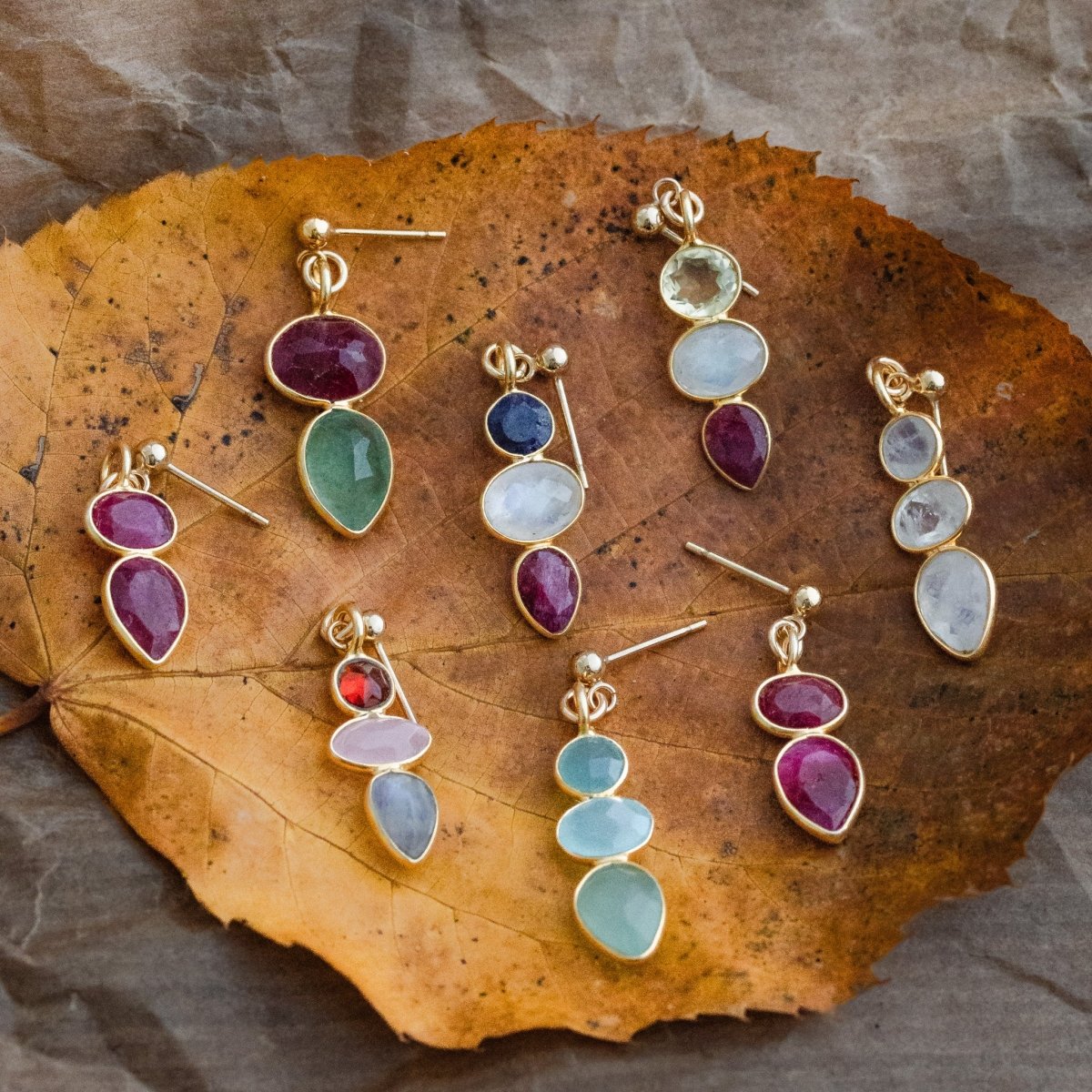 Acht bunte Multi-Edelstein Ohrstecker in gold liegen auf einem herbstlich-goldenen Blatt.